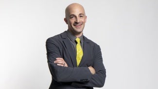 Headshot of a bald man in a dark jacket with a yellow tie standing in front of a gray background