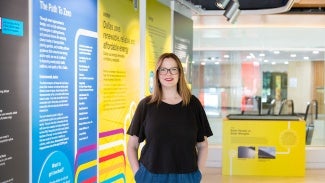 Woman with long hair and glasses in a black t-shirt stands in front on an exhibit.