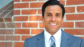 Headshot of a man with dark hair, wearing a gray suit and standing in front of a brick wall. 