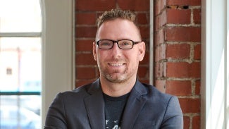 Headshot of a man wearing glasses and a blazer over a t-shirt.