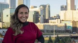 a woman with long hair and a red shirt stands in front of a cityscape.