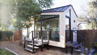 A tiny house with large glass windows, sits in the backyard, surrounded by a wooden fence and trees