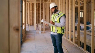 Man wearing PPE stands in wooden-framed construction site, looking at his clipboard