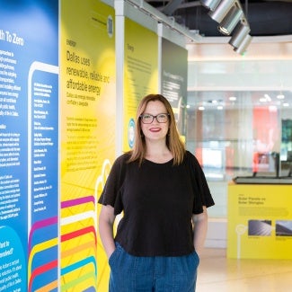 Woman with long hair and glasses in a black t-shirt stands in front on an exhibit.